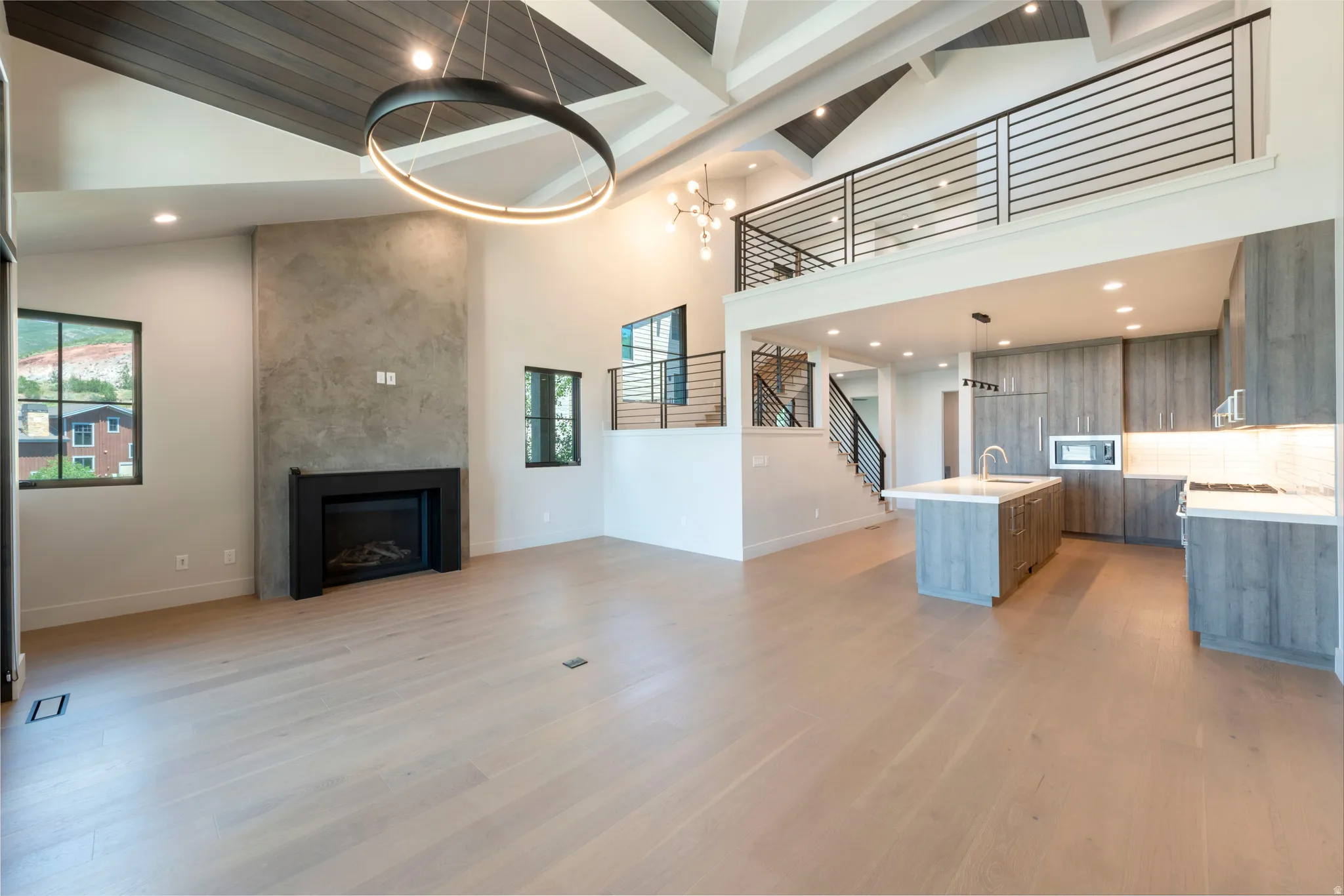Vaulted ceiling with exposed beams, wood-finish flooring, built-in fireplace with concrete surround, kitchen with wood-finish cabinetry and white countertops, and a mezzanine with metal railings