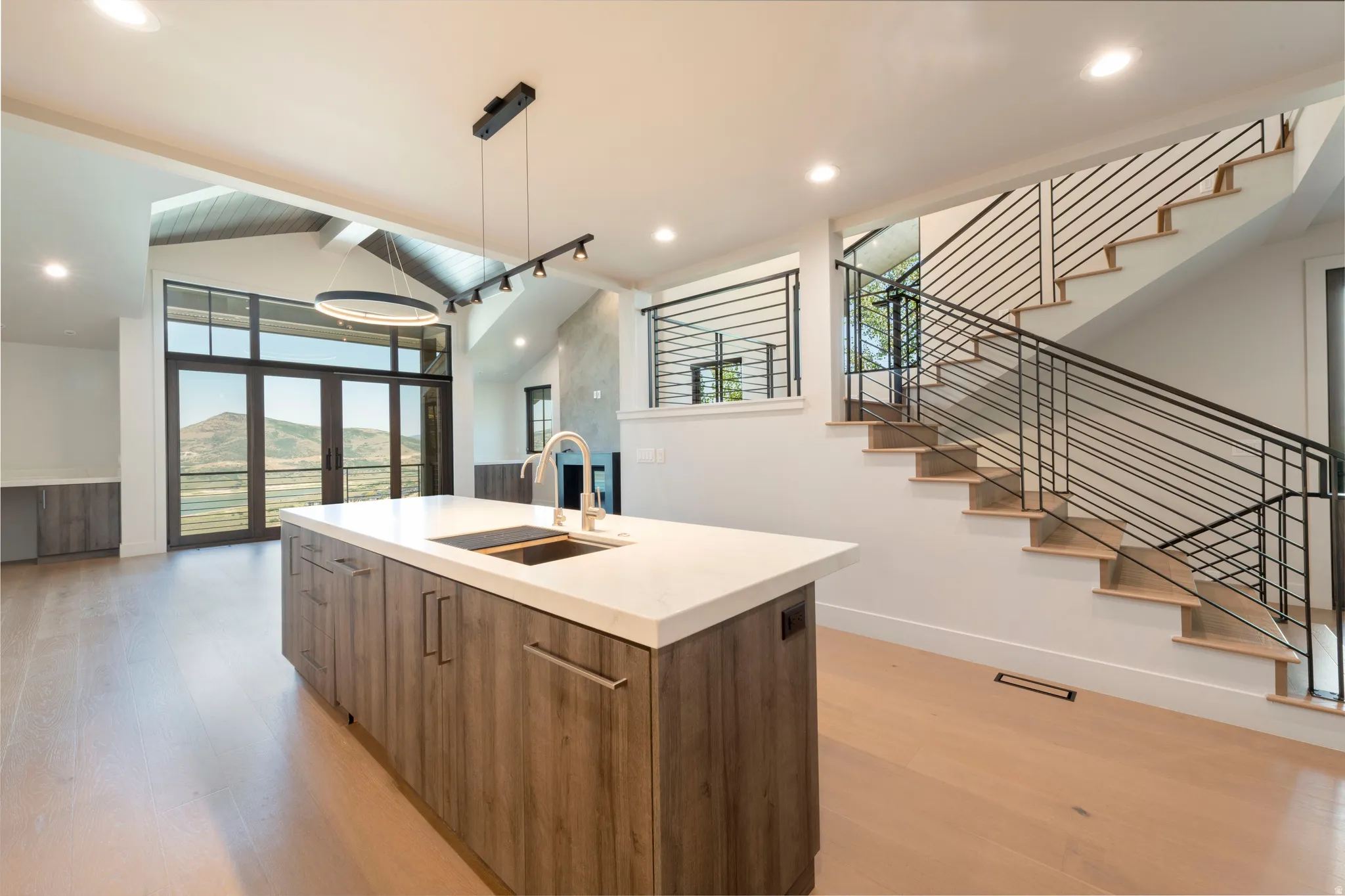 Kitchen island with a light-toned countertop and wood-finish cabinetry, light wood-finish flooring, staircase with horizontal metal railings, vaulted ceiling, and full-height windows