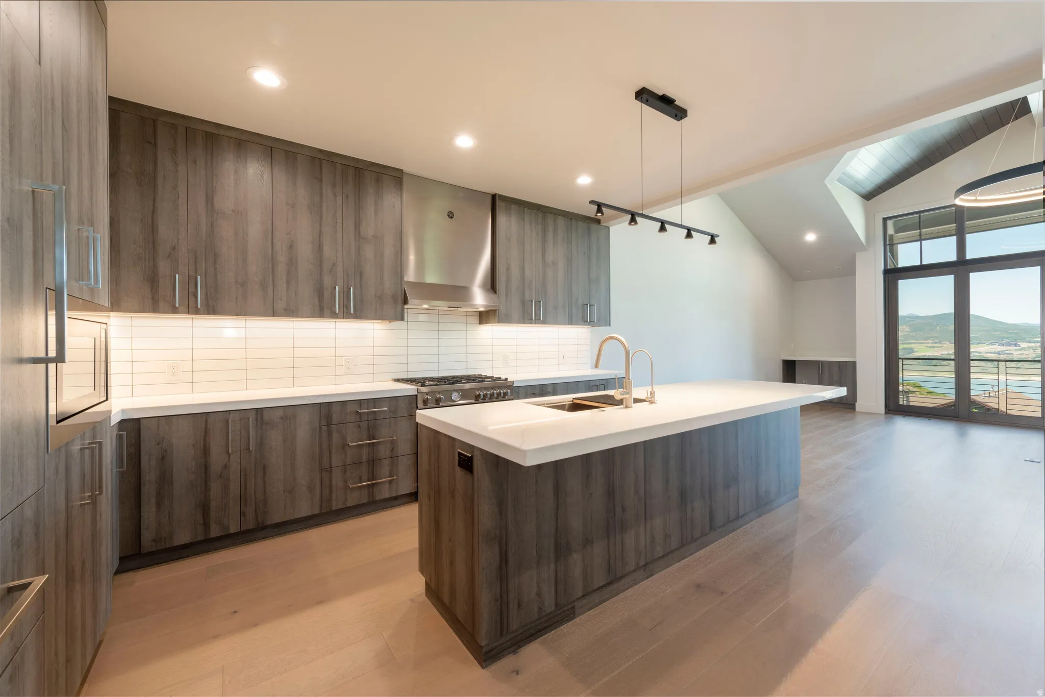 Modern kitchen featuring wood-finish cabinetry, light-toned countertops, a stainless steel range hood, and a large central island with a built-in sink