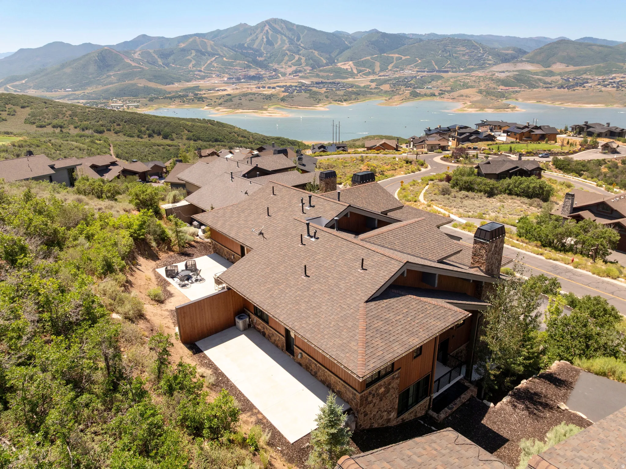 Expansive property featuring a brown shingle roof, stone and wood-paneled exterior, a concrete patio, and a mountain and lake backdrop