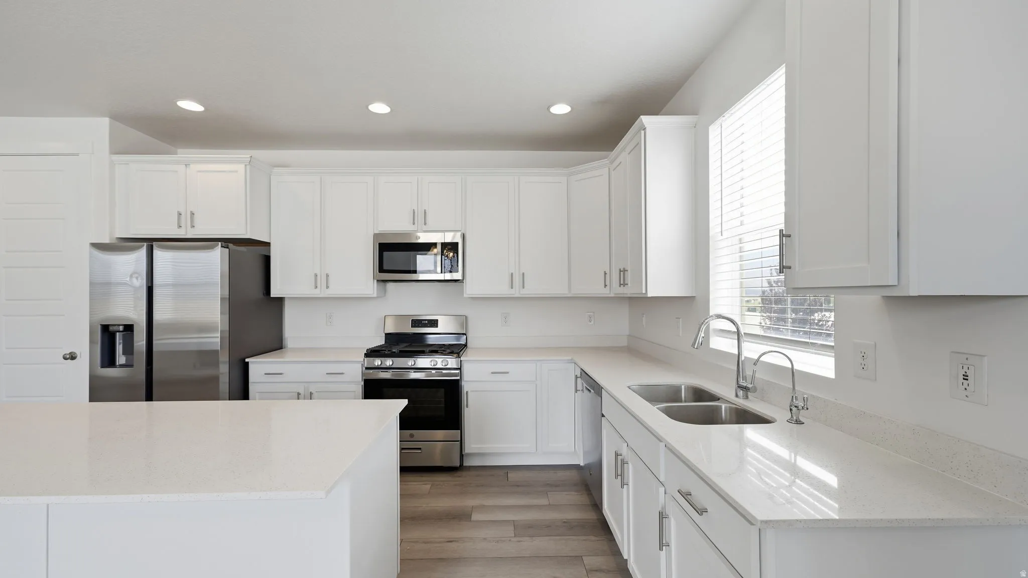 White shaker cabinetry, stainless steel appliances, light-toned countertops, wood-finish flooring, and recessed lighting