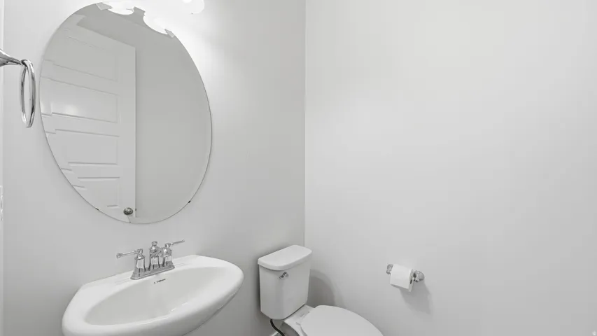Pedestal sink with chrome faucet, oval mirror, wall-mounted light fixture, and white toilet, off mud room