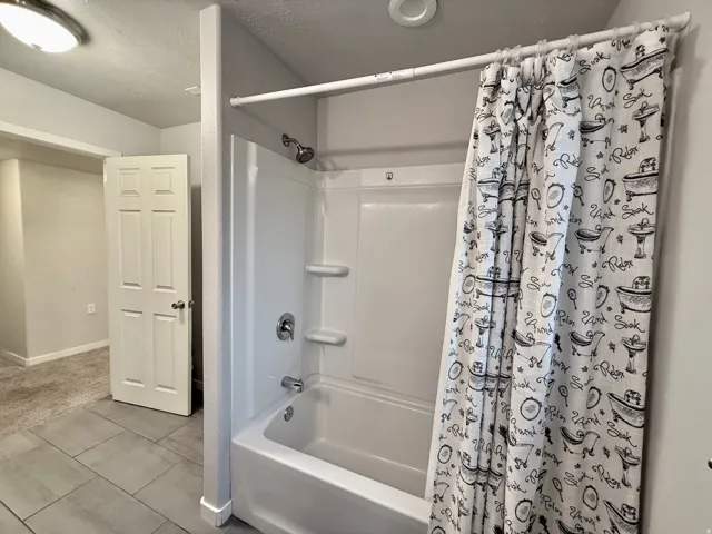 White fiberglass tub and shower combination with chrome fixtures, tiled flooring, and a flush mount ceiling light