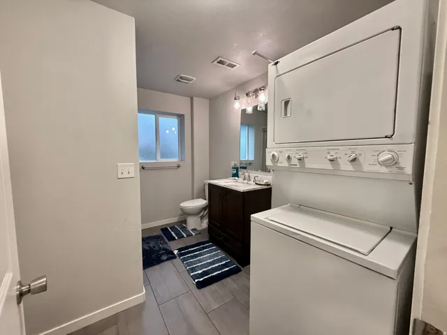 White stacked washer and dryer unit, dark wood-finish vanity with countertop, wall-mounted mirror with vanity lighting, white toilet, and gray tile flooring
