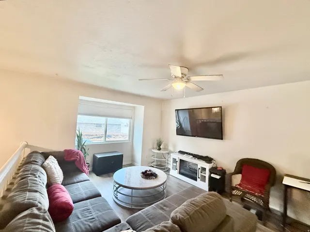 Wall-mounted television, built-in electric fireplace, wood-finish flooring, ceiling fan, and window with blinds