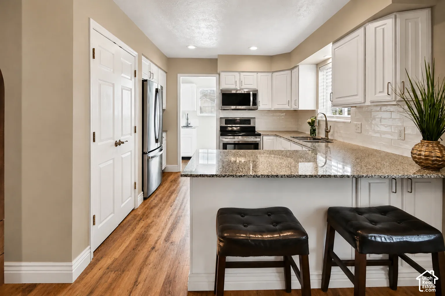 Stainless steel appliances, light-toned cabinetry, wood-finish flooring, a tile backsplash, and stone countertops