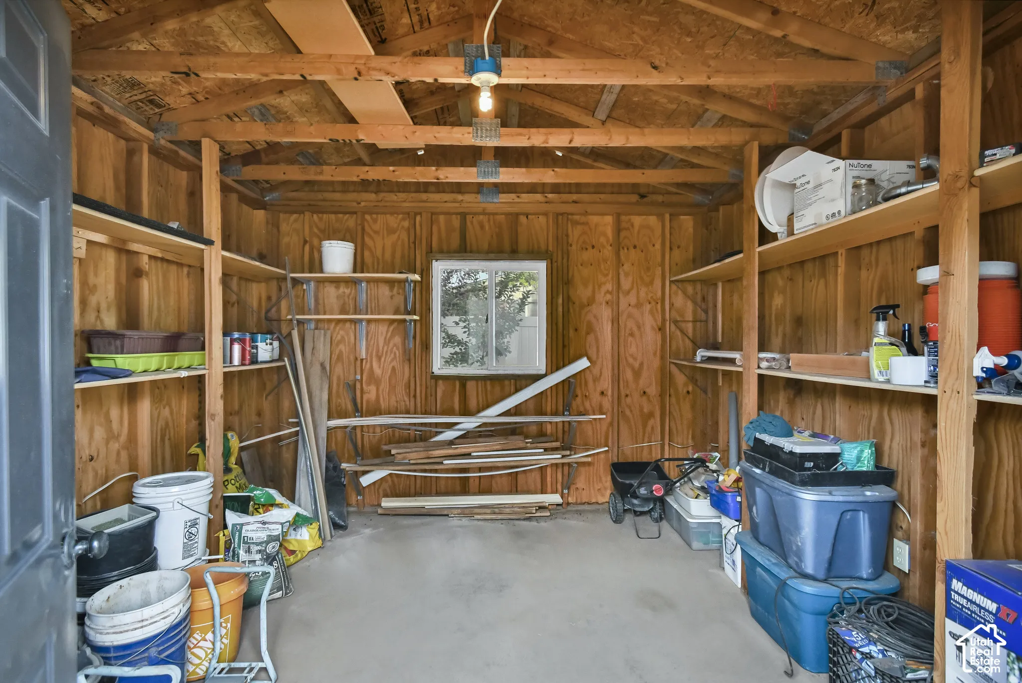 Wood-paneled interior with exposed beam ceiling, multiple built-in shelving units, concrete flooring, and a single window