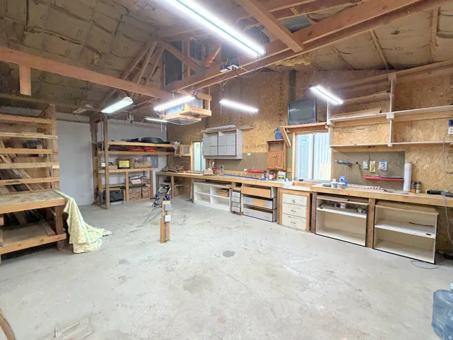 Concrete flooring, exposed wood beam ceiling, multiple fluorescent light fixtures, extensive built-in wooden shelving, and a large workbench with integrated cabinetry