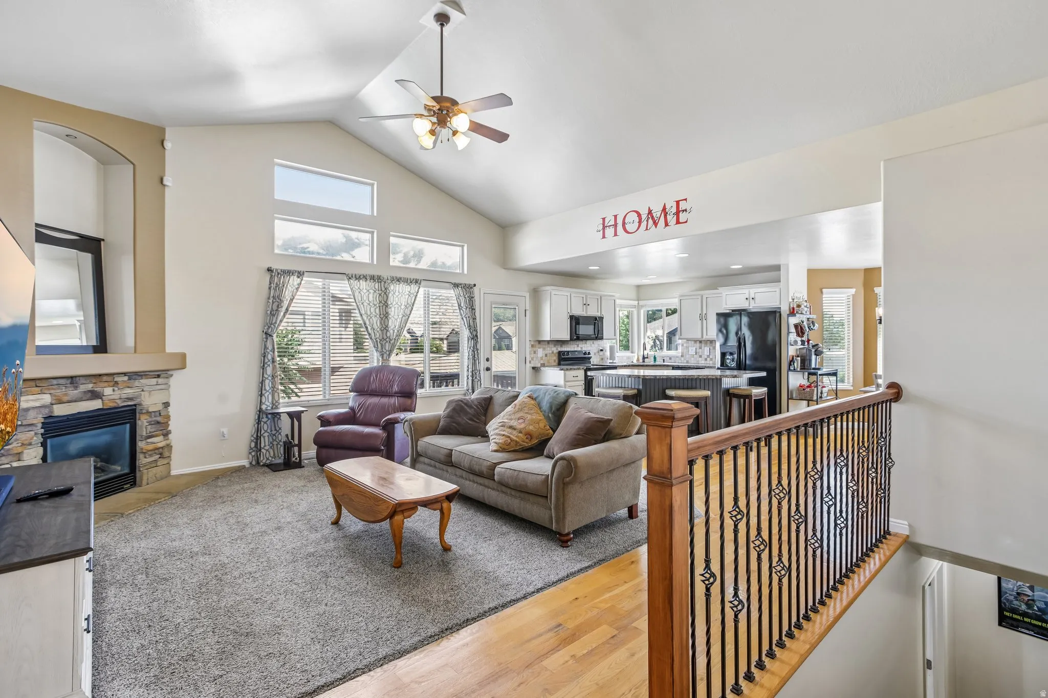 Vaulted ceiling with a ceiling fan, stone-surround fireplace, large windows, wood-finish flooring, and an open concept to the kitchen