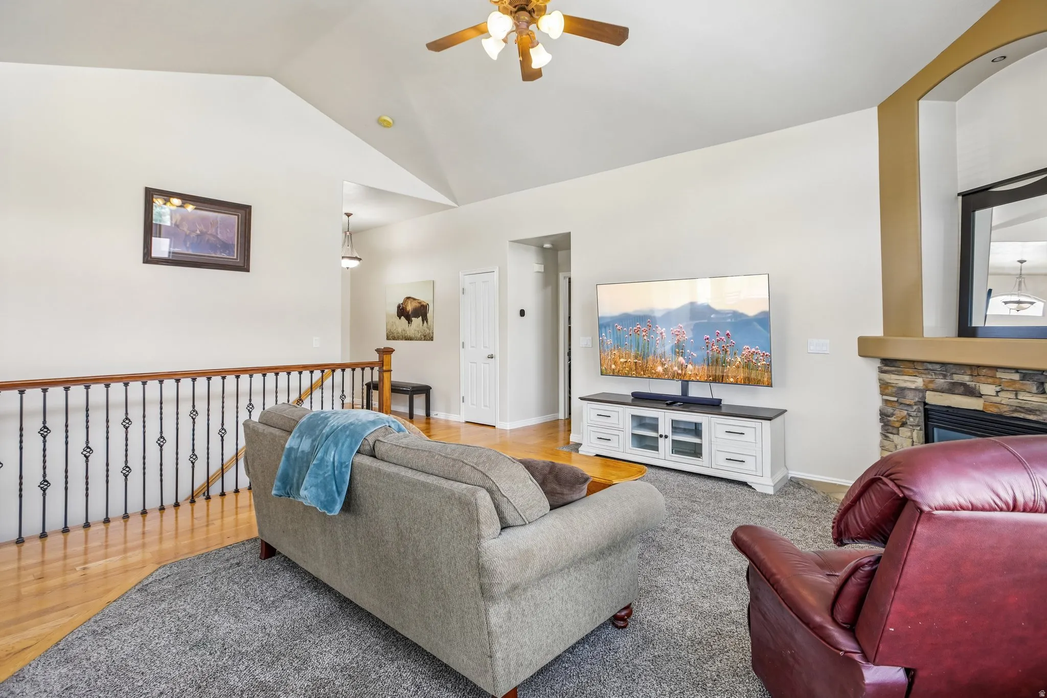 Vaulted ceiling with an attached ceiling fan, stone-clad fireplace, and a wood-finish railing with decorative metal spindles