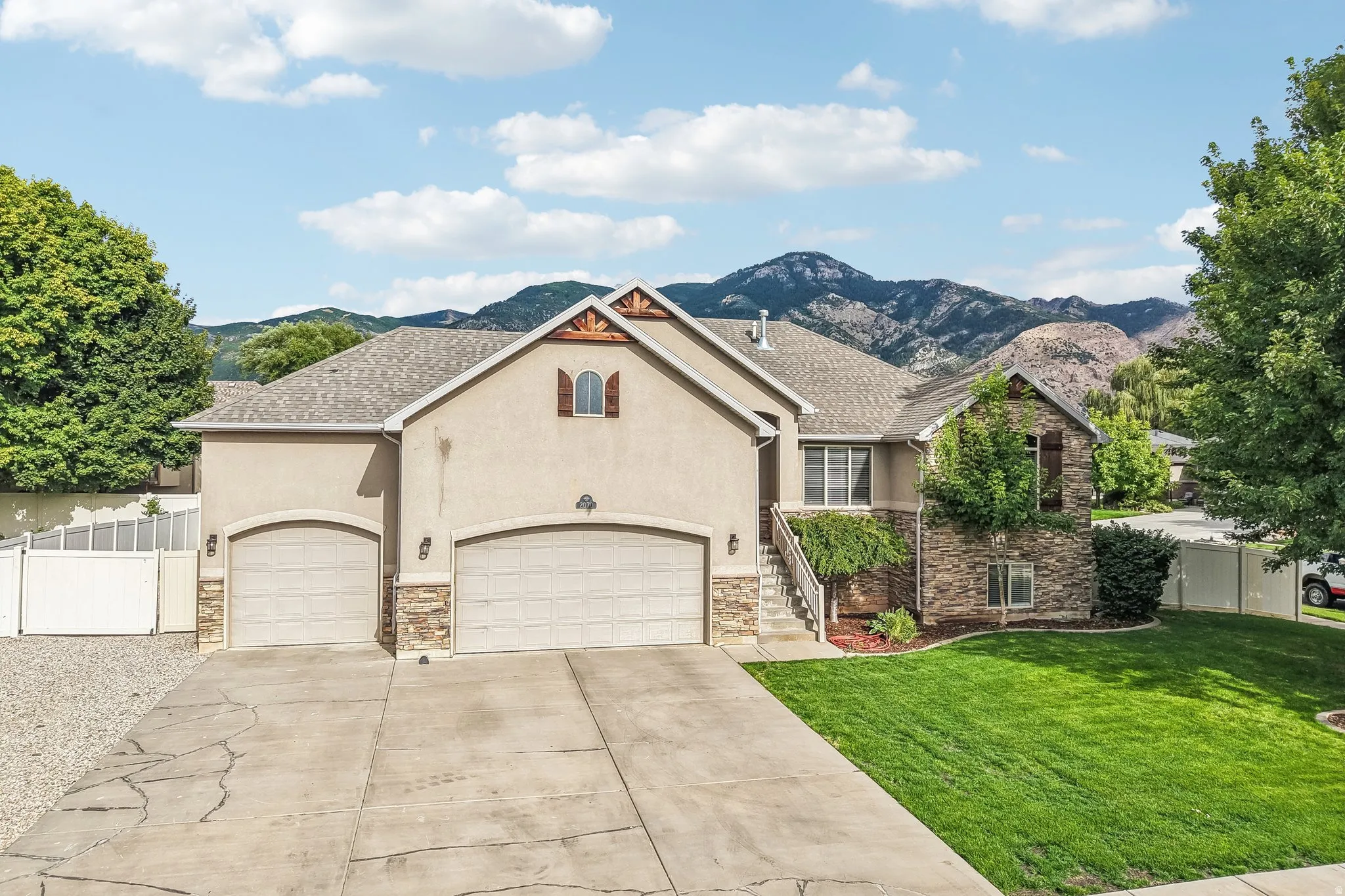 Stucco and stone exterior with architectural wood details, two-car garage, mature front landscaping, and a concrete driveway