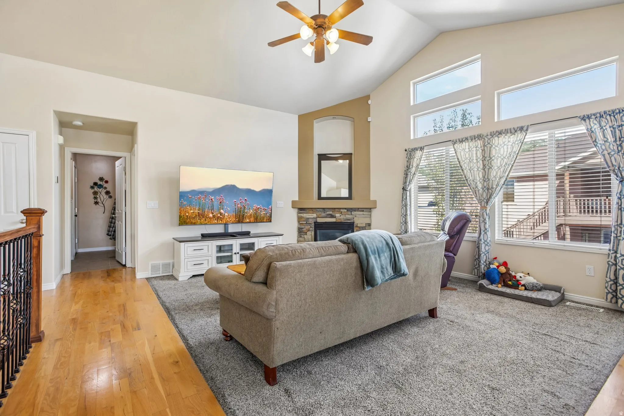 Vaulted ceiling with a ceiling fan, stone-clad fireplace, hardwood flooring, and large windows