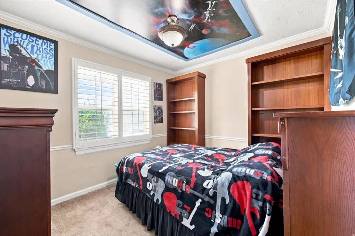 Built-in bookcases, window with plantation shutters, crown molding, and recessed ceiling with decorative mural