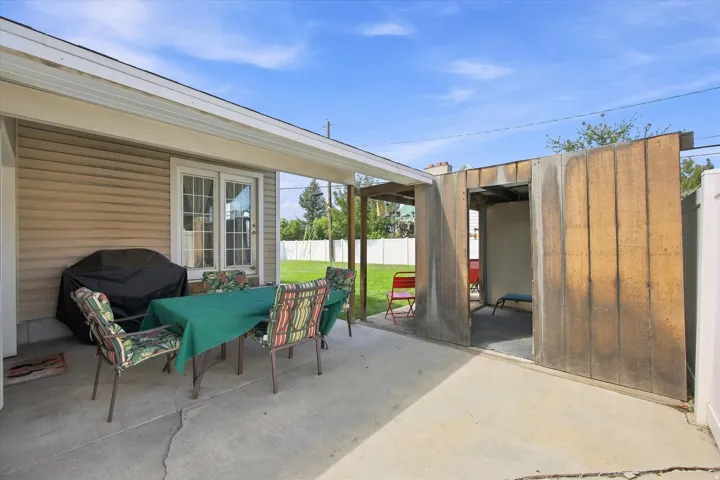 Covered patio with concrete flooring, framed by a white fence and a detached wood-sided structure