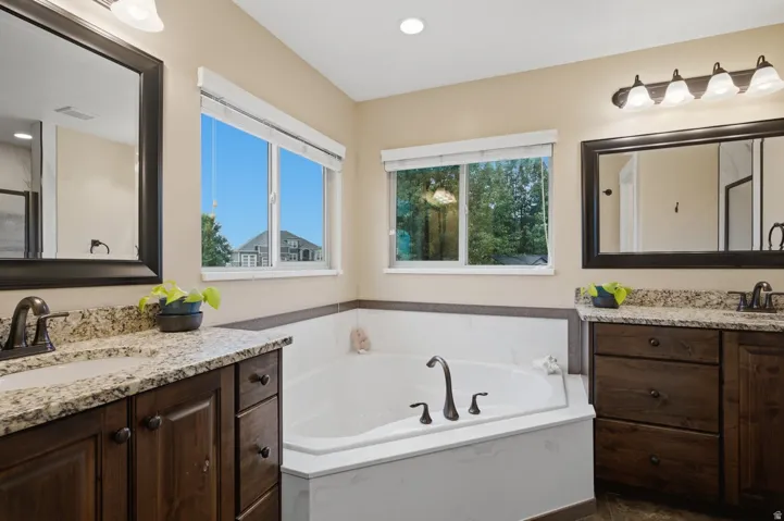 Dual wood-finish vanities with light-toned countertops, an integrated corner soaking tub, and natural light from multiple windows