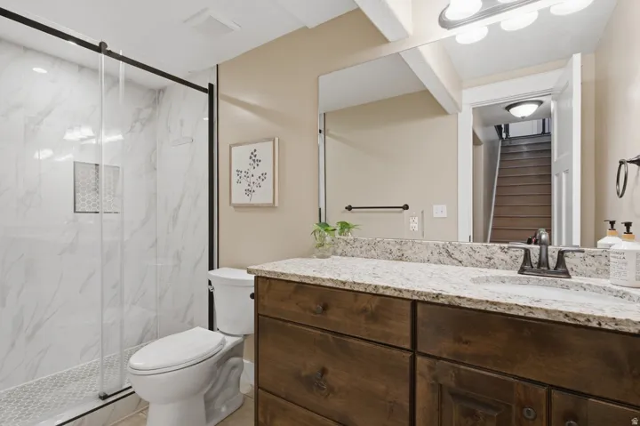 Walk-in shower with clear glass enclosure and hexagonal tile flooring, wood-finish vanity with stone countertop, and an undermount sink
