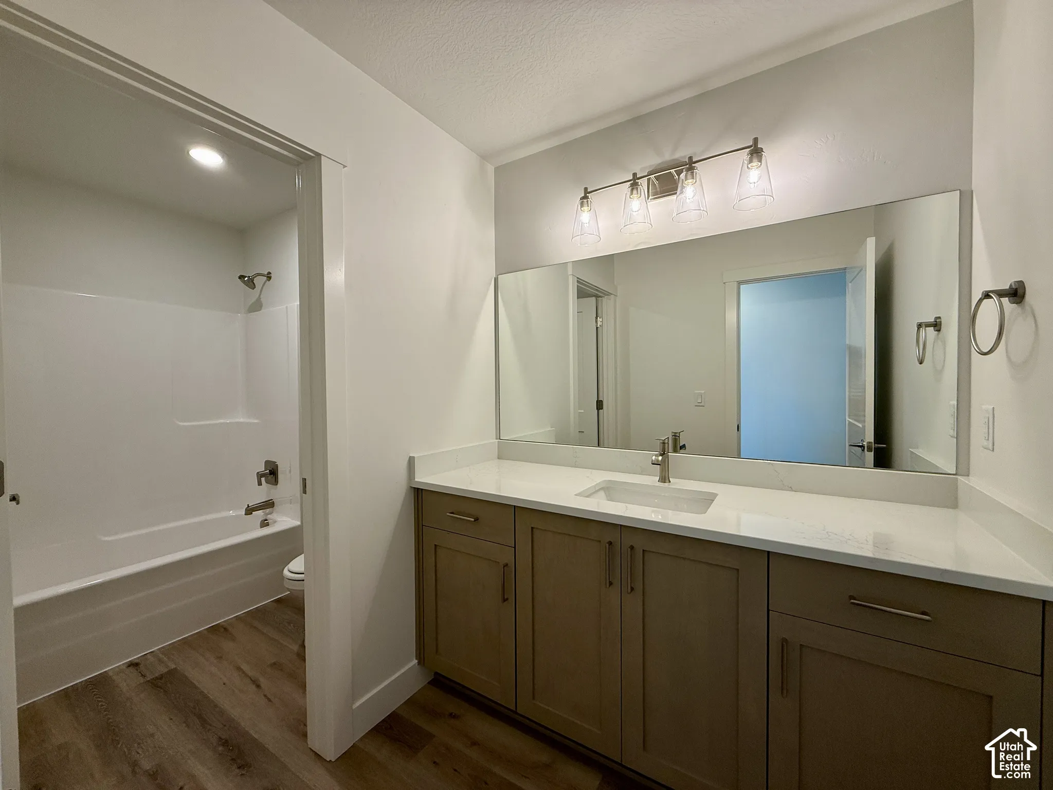 Bathroom featuring a vanity with shaker-style cabinetry and a stone countertop, an integrated rectangular sink, a full-width vanity mirror, a four-light fixture, and a tub-shower combination with wood-finish flooring