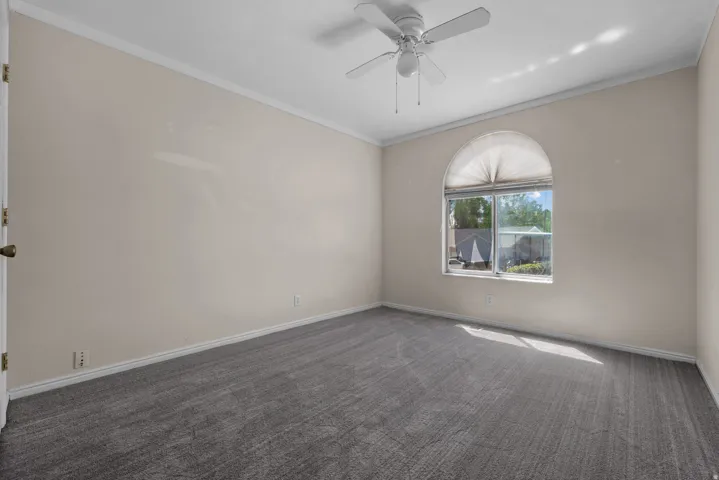 Neutral-toned walls with crown molding, gray patterned carpeting, arched window with pleated shade, and a ceiling fan with light fixture