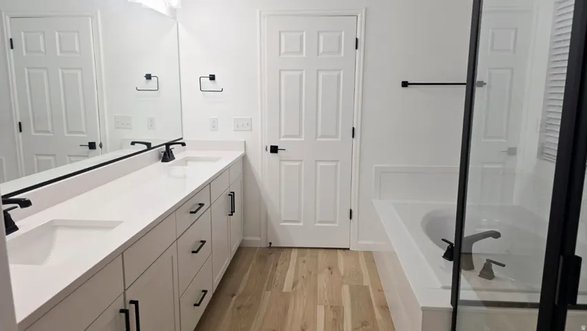 Dual vanity with undermount sinks and matte black fixtures, extensive countertop space, built-in soaking tub, glass-enclosed shower, and wood-finish flooring