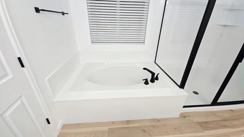 Built-in soaking tub with matte black fixtures, framed glass shower, light wood-finish flooring, and a window with blinds