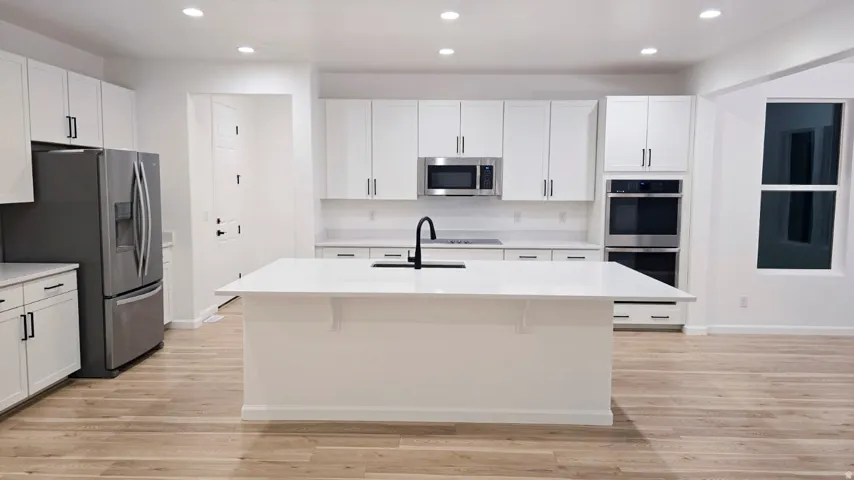 Modern kitchen featuring white cabinetry, a large center island with a black faucet, recessed lighting, stainless steel appliances, and light wood-finish flooring