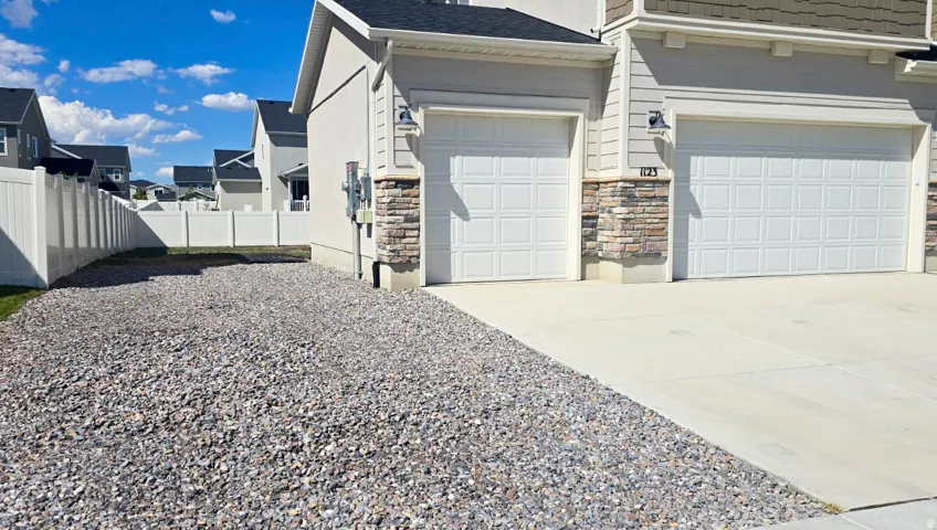 Light-colored siding with stone accent, concrete driveway, two garage doors, outdoor lighting fixtures, and a gravel side yard