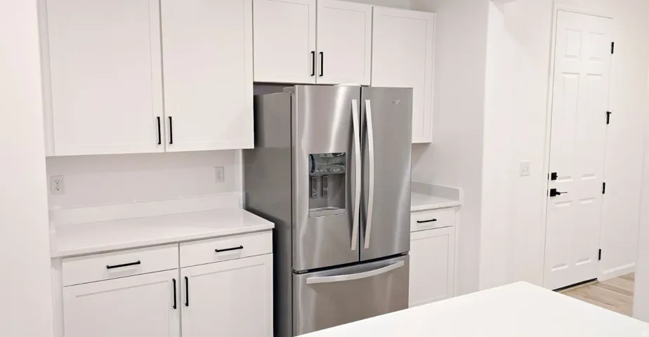 White shaker cabinetry, stainless steel refrigerator, white countertops, panel door with black hardware, and light wood-finish flooring