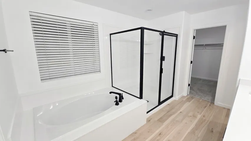 Integrated soaking tub with matte black fixtures, separate enclosed shower with black framing, wood-finish flooring, and a window with horizontal blinds