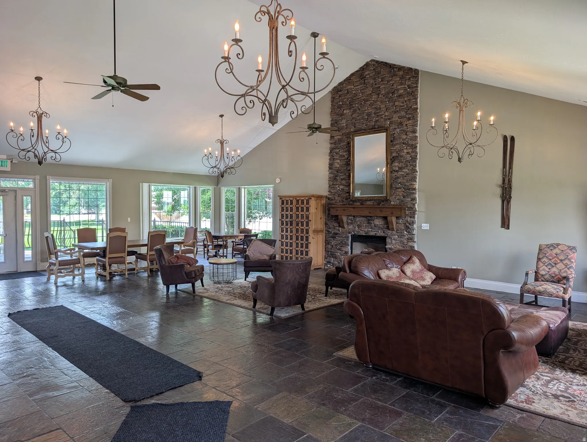 Spacious common area featuring a vaulted ceiling with exposed beams, multiple decorative chandeliers, a stone-clad fireplace with a wood mantle, and large windows