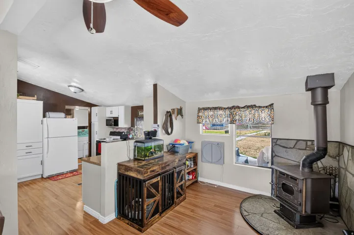 Open-concept kitchen and living area featuring wood-finish flooring, white cabinetry with ample storage, a central island with shelving, stainless steel appliances, a wood-burning stove with black metal surround, and a ceiling fan
