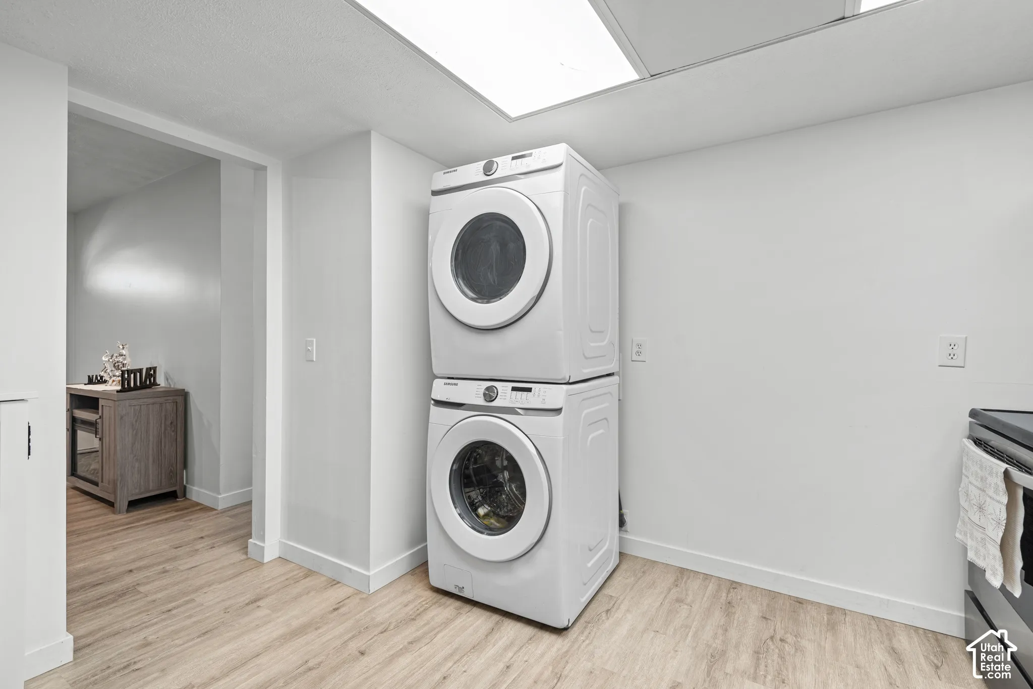Stacked laundry appliances, overhead fluorescent lighting, wood-finish flooring, and white walls