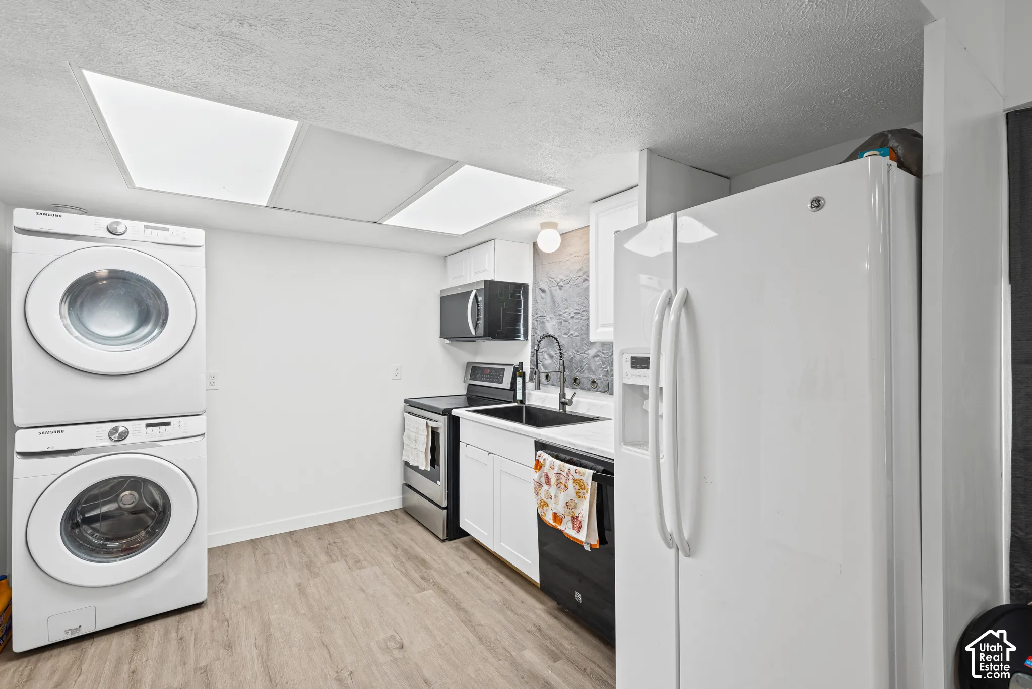 Integrated stacked washer and dryer, wood-finish flooring, white cabinetry, dark gray textured backsplash, and a black sink with a matte black faucet