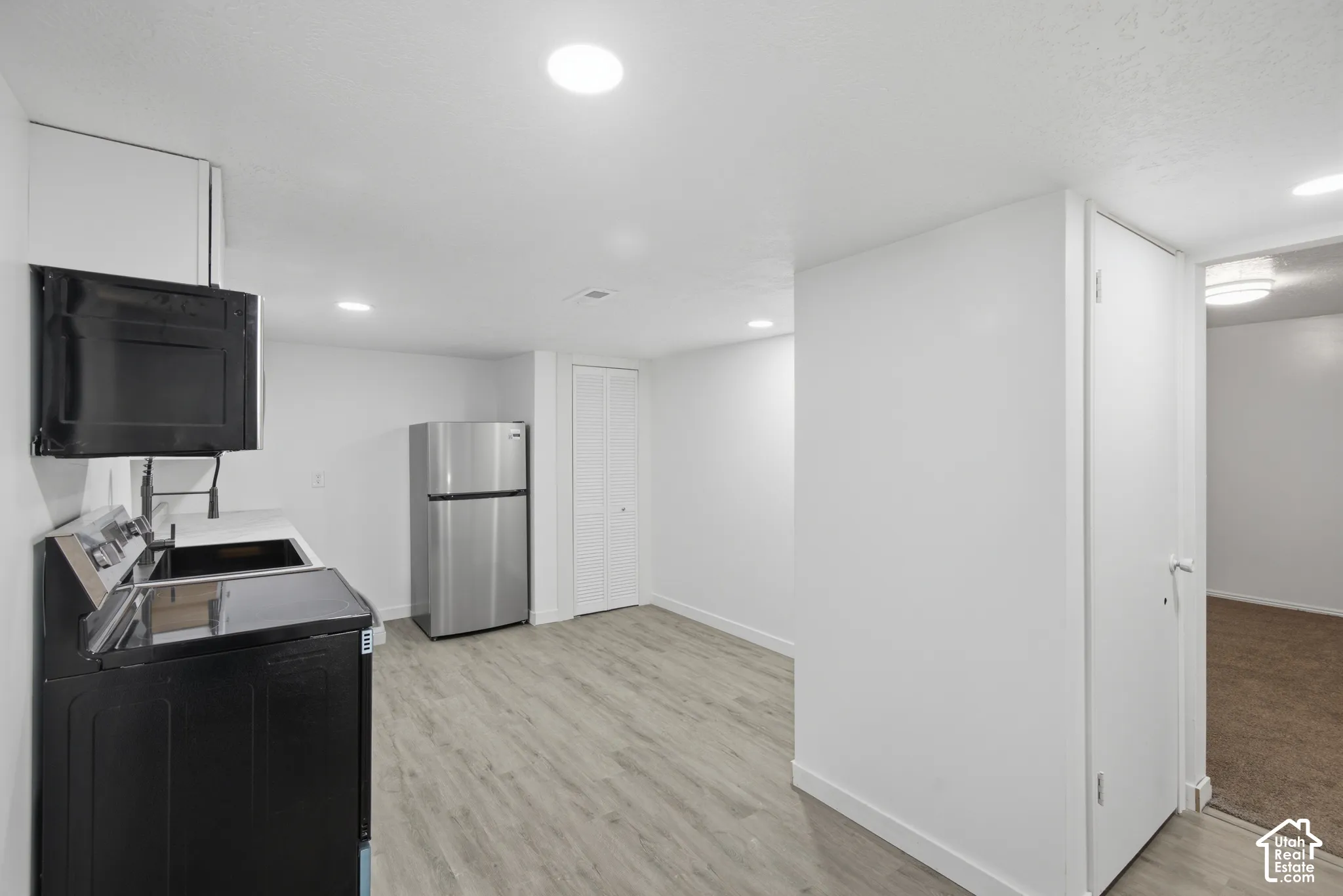 Recessed lighting throughout, light wood-finish flooring, stainless steel refrigerator, white walls, and a built-in microwave