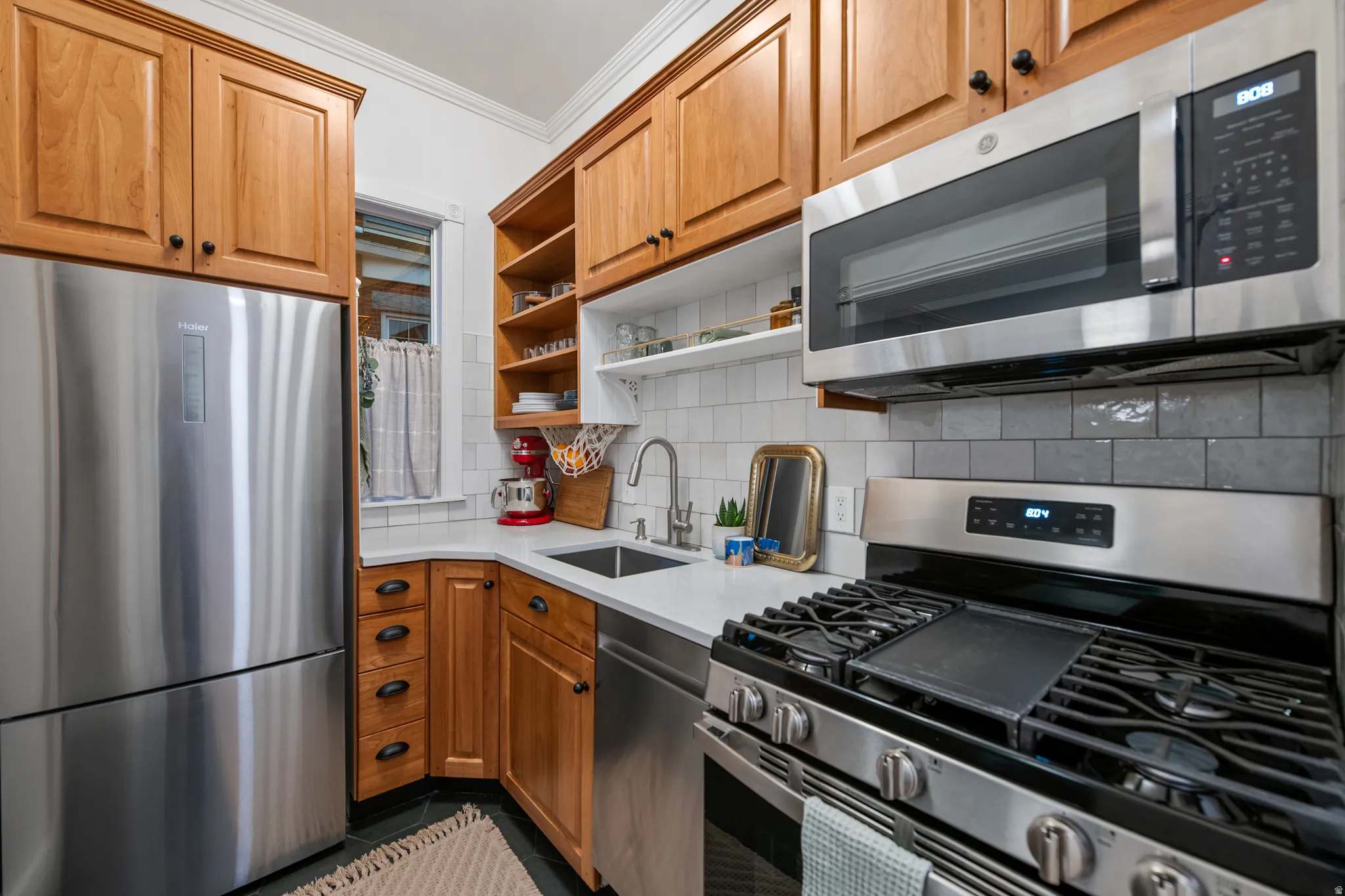 Stainless steel appliances, wood cabinetry, built-in shelving, white subway tile backsplash, and light-toned countertops