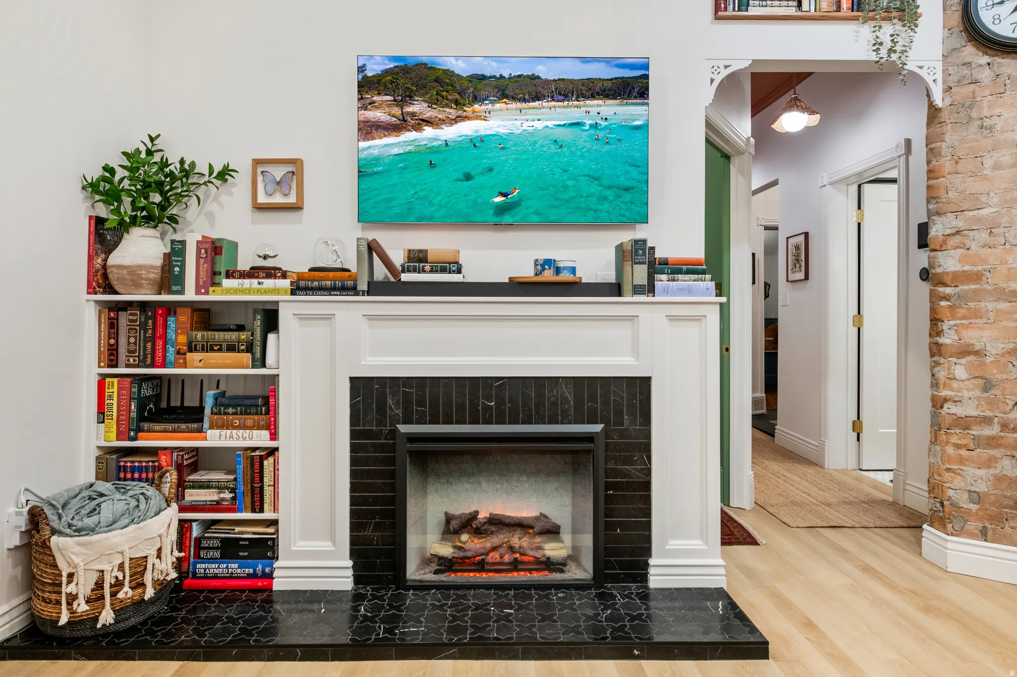 Built-in shelving unit, white mantel with dark tile surround, integrated fireplace, light wood-finish flooring, and exposed brick wall