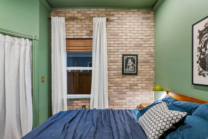 Exposed brick accent wall, double-hung window with wood-finish blinds, light green wall paint, coordinating green ceiling, and white curtains