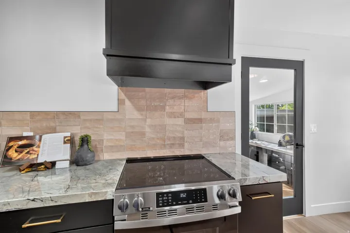 Stainless steel electric range, stone countertops, tiled backsplash, dark wood cabinetry, and a dark-framed glass paneled door