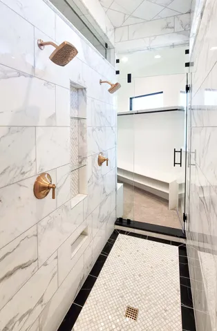 Walk-in shower with large format marble-look tile, dual copper-finish shower heads, integrated wall niches, and a mosaic tile floor with a contrasting border