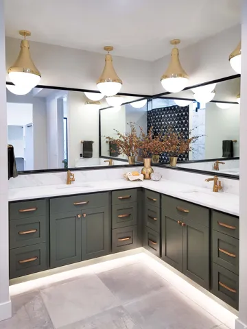 Luxurious primary bathroom featuring a corner dual-vanity setup with white quartz countertops, sage green cabinetry with brass hardware, and an expansive framed mirror