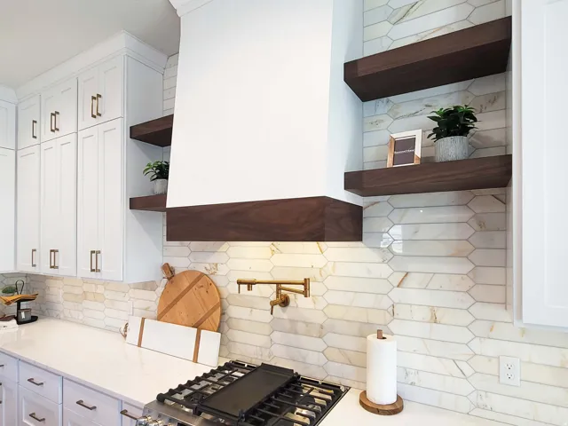White shaker cabinetry with brass hardware, wood-finish floating shelves, white range hood with wood-finish trim, gas range, and a tiled backsplash
