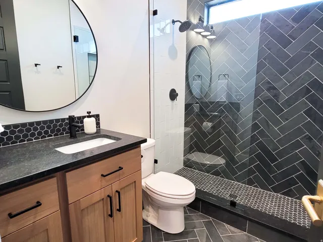 Bathroom featuring a clear glass shower enclosure, dark herringbone tile shower surround, honeycomb tile shower floor, wood-finish vanity with dark countertop, and hexagonal tile backsplash