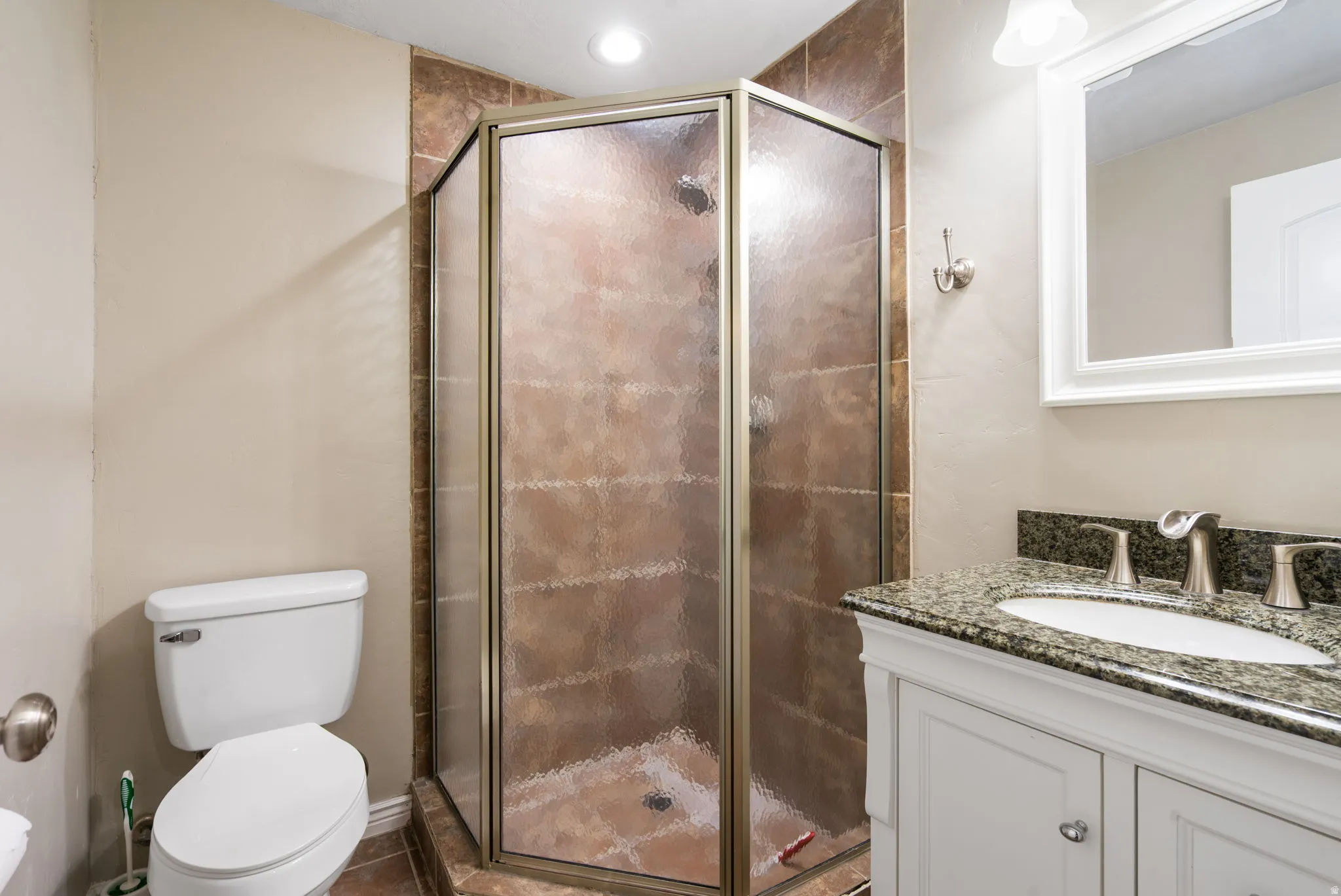 Spa-inspired ensuite bathroom featuring a corner shower enclosure with bronze-framed glass doors and earth-tone tile surround, paired with a white vanity topped in speckled quartz countertop, polished chrome fixtures, and recessed ceiling lighting