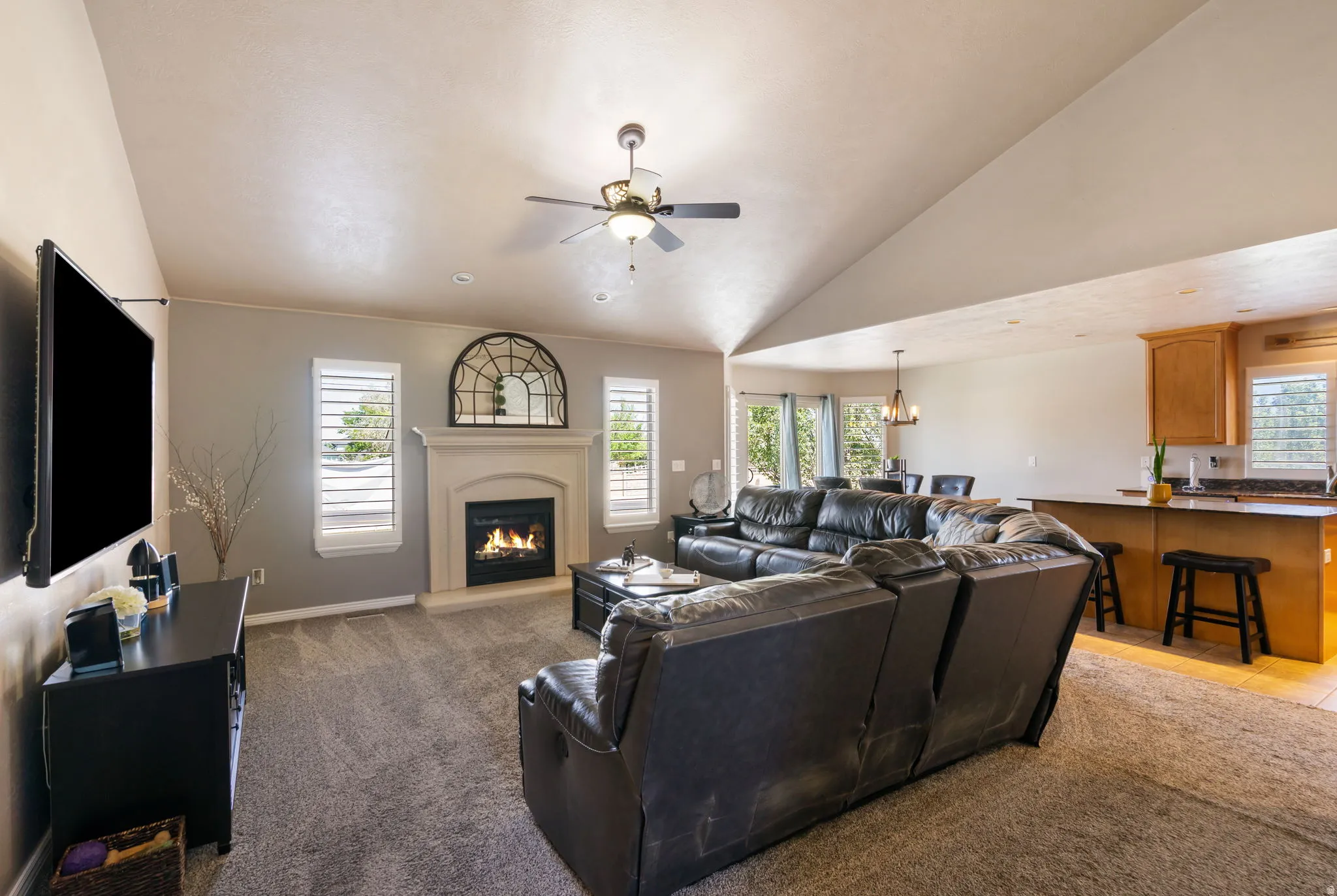 Vaulted ceiling, ceiling fan, fireplace with mantel, window shutters, and kitchen with wood-finish cabinetry