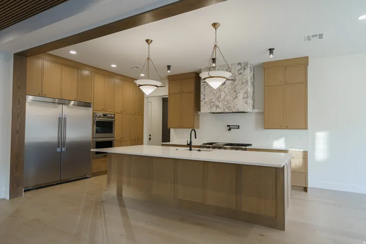 Stainless steel appliances, built-in double ovens, light wood-finish cabinetry, a large kitchen island with a light-colored countertop, and a decorative range hood