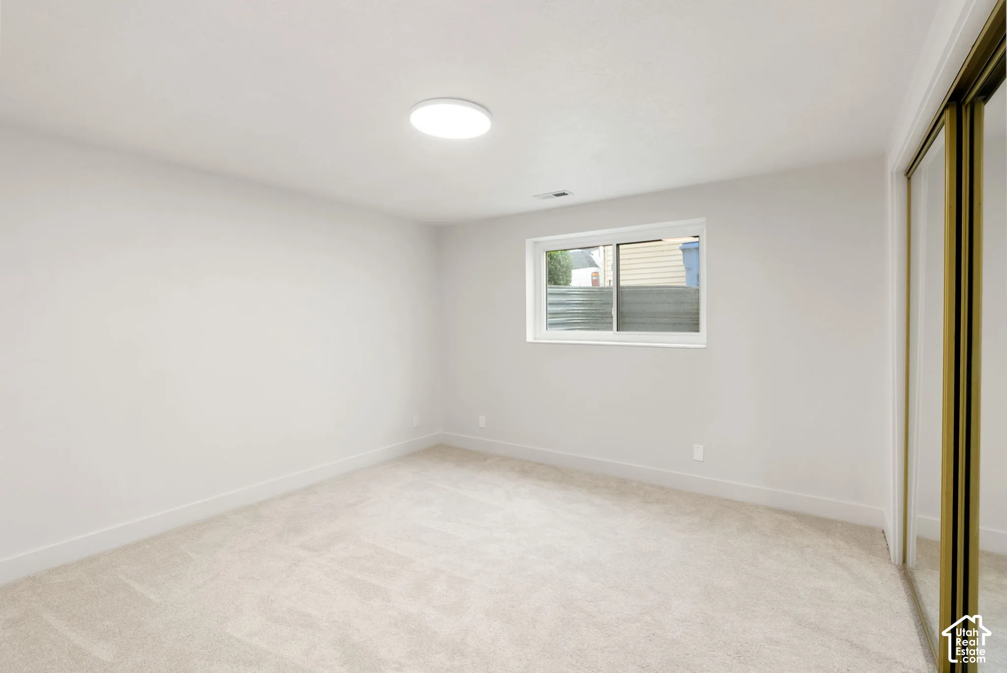 Neutral-toned wall-to-wall carpeting, white painted walls, a flush-mount ceiling light fixture, a window with a white frame, and a mirrored sliding closet door