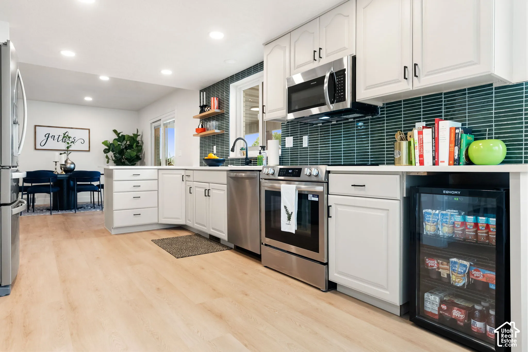 Modern kitchen featuring light wood-finish flooring, white cabinetry, stainless steel appliances, a beverage cooler, and a teal tile backsplash