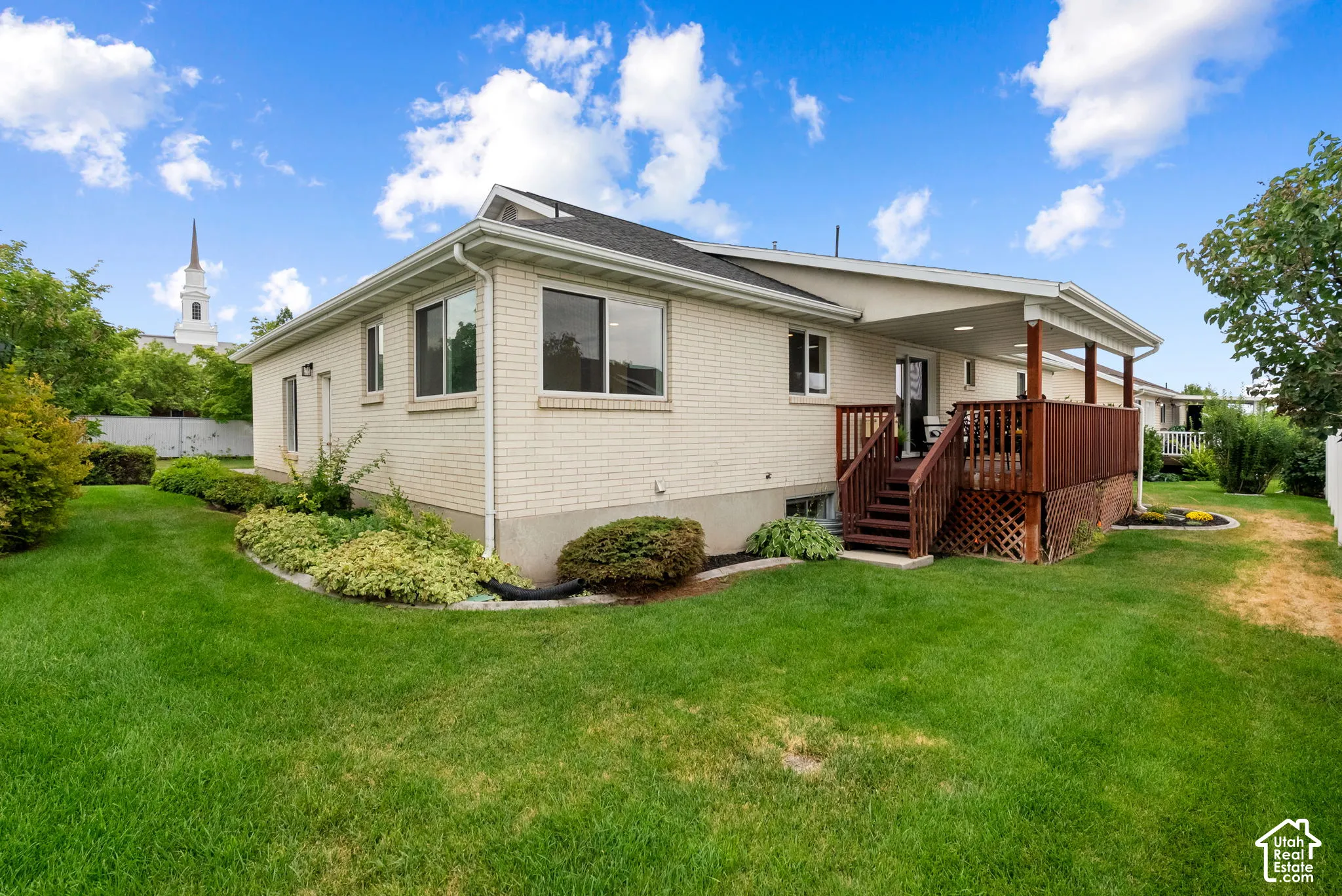 Brick exterior with white trim, covered wooden deck, mature landscaping, and a manicured lawn