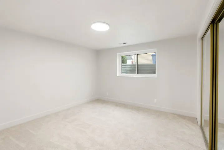 Neutral-toned wall-to-wall carpeting, white painted walls, a flush-mount ceiling light fixture, a window with a white frame, and a mirrored sliding closet door