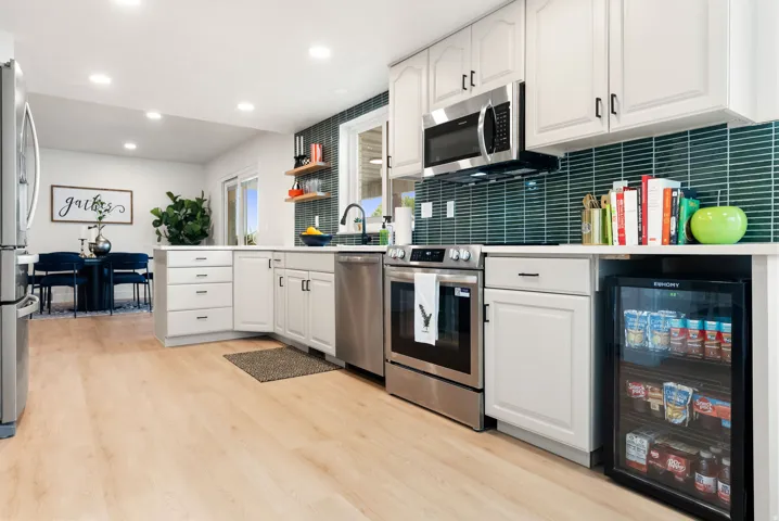 Modern kitchen featuring light wood-finish flooring, white cabinetry, stainless steel appliances, a beverage cooler, and a teal tile backsplash