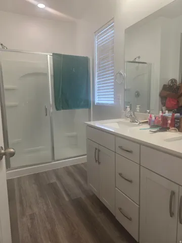 Dual vanity with white cabinetry, wood-finish flooring, and a shower with a sliding glass enclosure